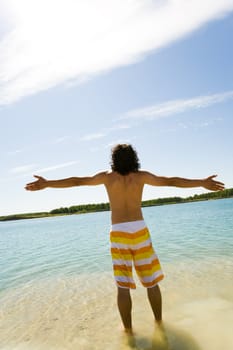Rear view of young man standing in blue water and having fun with outstretched arms on hot summer day