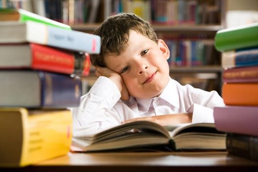 Portrait of cheerful lad sitting in library before book and looking at camera with smile