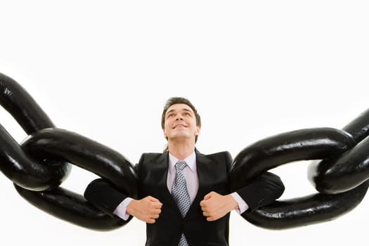 Portrait of powerful businessman holding two sections of huge chain by arms and looking upwards with smile over white background