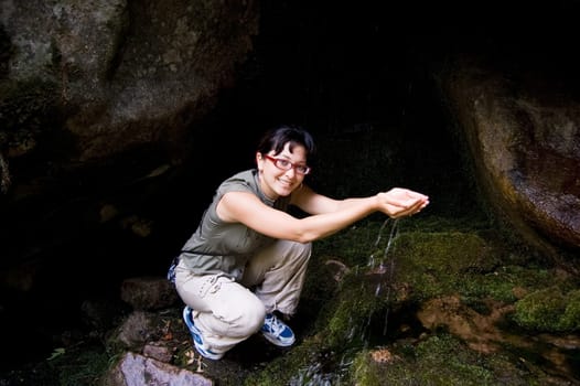 Photo of happy female putting her hands under water splashes in the mountains and looking at camera with smile