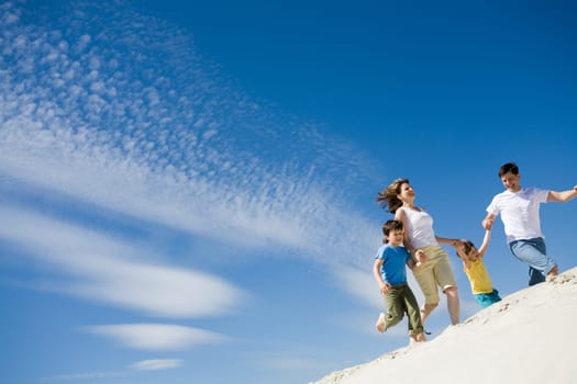 Photo of happy parents holding their sons by hands and running down sandy shore with bright blue sky at background