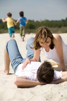 Image of amorous couple lying on sandy beach with their sons at background