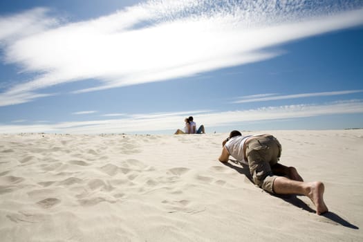 Careful photograpther taking shots of relaxing couple sitting back to back on sandy beach against blue sky