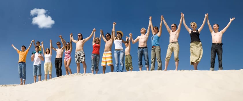Image of many friends standing on sandy beach with their arms raised against blue sky