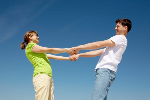 Photo of playful couple holding each other by hands with their arms stretched against blue sky