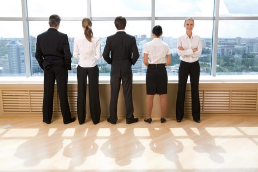 Rear view of business group standing in row and looking through window with their leader smiling at camera