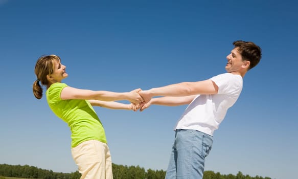 Photo of playful couple holding each other by hands with their arms stretched against blue sky
