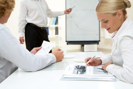 Photo of pretty student making notes at workplace on background of teacher by the whiteboard
