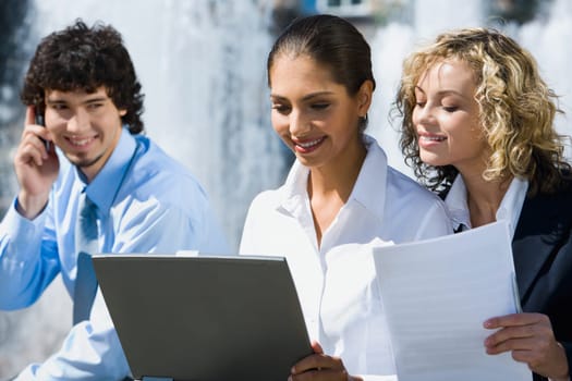 Two cute smiling female students are reading documents outside and handsome young man talking by the phone staring at them