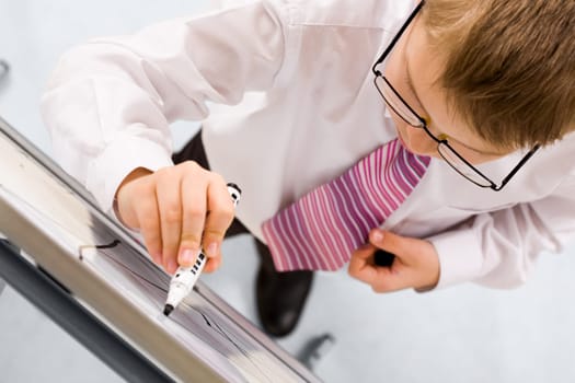 Smart boy with pen in hand standing by whiteboard and writing on it