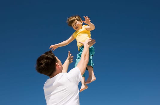 Photo of happy man throwing his cute son up with bright blue sky at background