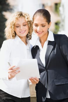 Two cute smiling female students standing outside ahead of the fountain reading the documents