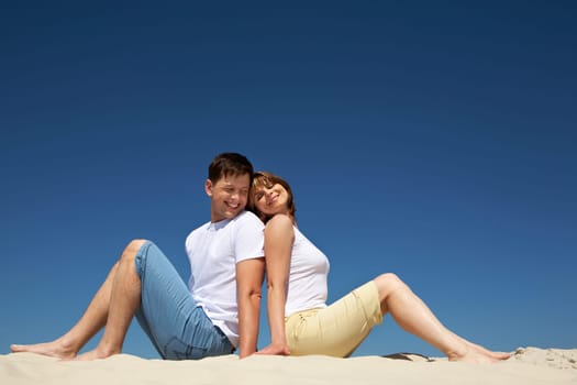 Portrait of relaxing couple sitting back to back on sandy beach against blue sky