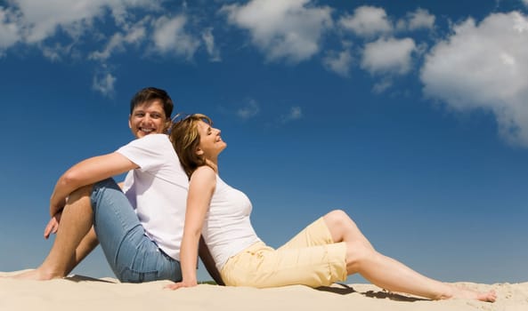 Portrait of relaxing couple sitting back to back on sandy beach against blue sky