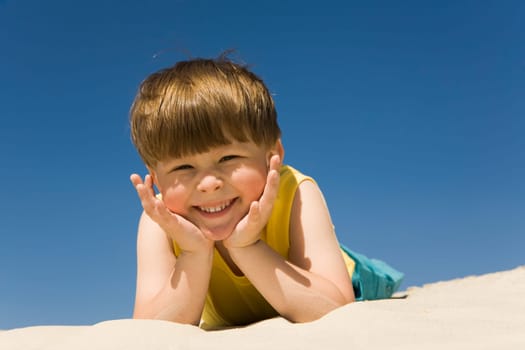 Happy boy lying on sand and looking at camera with smile on hot summer day