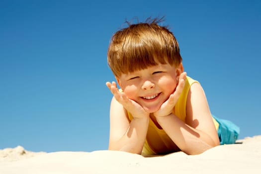 Happy boy lying on sand and looking at camera with smile on hot summer day