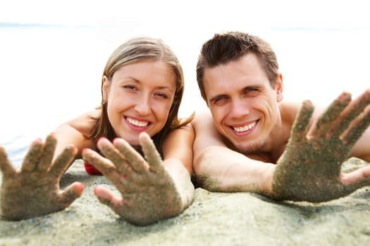 Photo of smiling couple lying on beach and showing their hands covered with sand while sunbathing