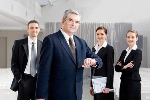 Portrait of senior businessman looking at camera while watching the time at background of confident managers