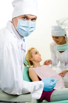 Portrait of confident doctor in sterile mask on background of his assistant helping patient