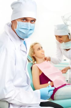 Portrait of confident doctor in sterile mask on background of his assistant helping patient