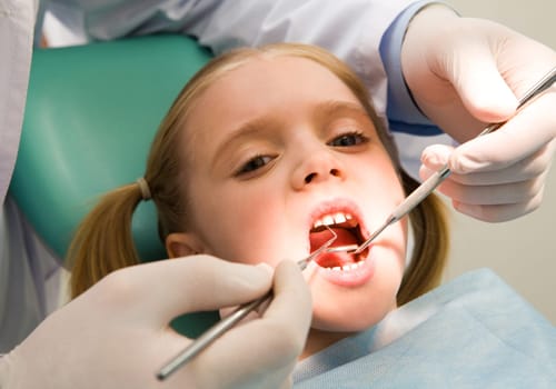 Photo of small girl looking at camera with open mouth while it being examined by dentist
