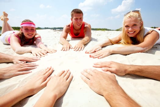 Image of several teens lying on beach and keeping their hands on sand in the form of circle