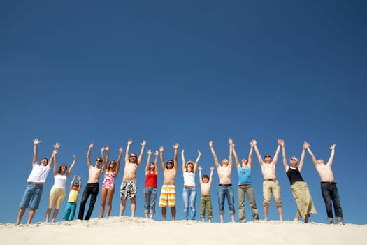 Image of many friends standing on sandy beach with their arms raised against blue sky