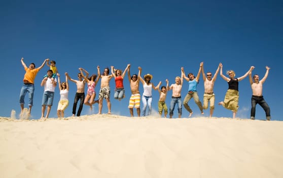 Crowd of friends jumping on sandy beach with their arms raised against blue sky