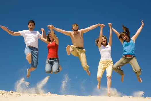 Photo of excited people jumping on sandy beach with their arms raised against blue sky