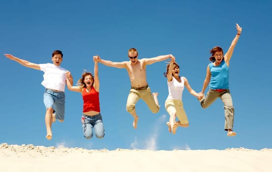 Photo of excited people jumping on sandy beach with their arms raised against blue sky