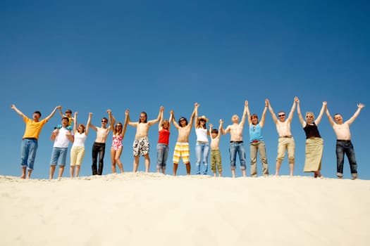 Image of many friends standing on sandy beach with their arms raised against blue sky