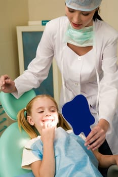 Photo of happy girl looking at her teeth in the mirror with helpful assistant near by