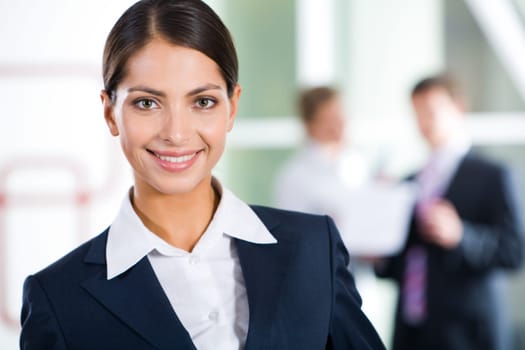 Portrait of clever businesswoman looking at camera in the room with two businesspeople on the background