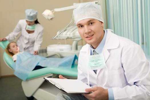 Image of handsome doctor with paper in hands looking at camera on background of assistant and little girl