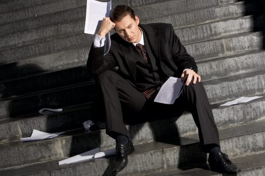 Portrait of sad businessman sitting on stairs with papers in hands and lost expression on his face