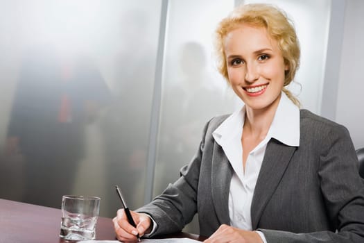 Portrait of pretty smiling woman writing and sitting at the table with glass of water, paper on it
