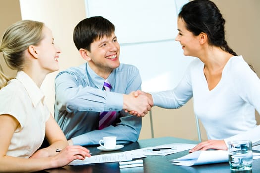 Image of business partners shaking hands at business meeting with a smiling businesswoman sitting near by