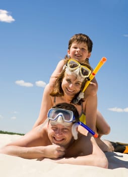 Portrait of happy family in goggles and flippers lying on sandy beach against blue sky