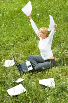 Portrait of happy woman with papers in hands sitting on green glade and raising her arms
