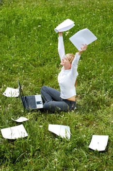 Portrait of happy woman with papers in hands sitting on green glade and raising her arms