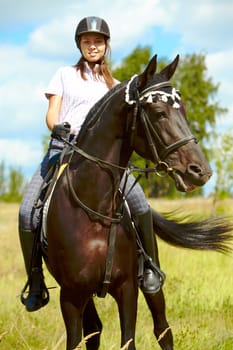 Image of happy female sitting on purebred horse and looking at camera outdoors