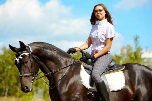 Image of happy female in sunglasses sitting on purebred horse outdoors