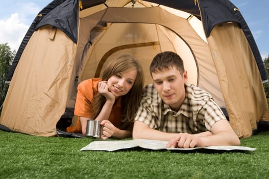 Photo of resting couple lying on green grass in tent and reading guide