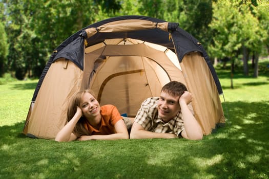 Photo of resting couple lying on green grass in tent and looking at camera with smiles