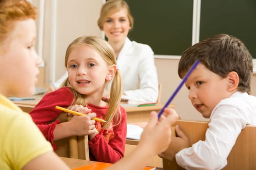 Portrait of schoolchildren sitting in classroom and speaking with their teacher behind