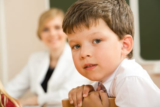 Face of clever schoolboy looking at camera in classroom with his teacher at background
