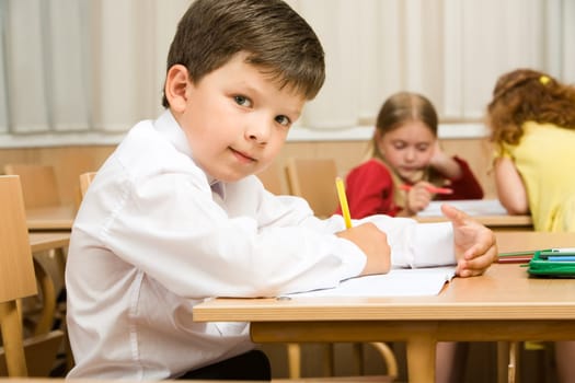 Image of smart schoolboy sitting at desk and drawing while looking at camera during lesson