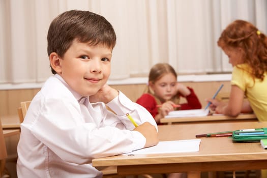 Image of smart schoolboy sitting at desk and looking at camera during lesson