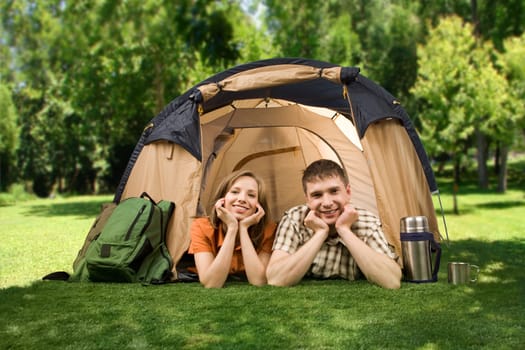 Photo of cheerful girl and guy lying on green grass in tent and looking at camera with smiles