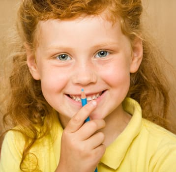 Face of red-headed girl holding blue pencil by her mouth looking at camera with smile